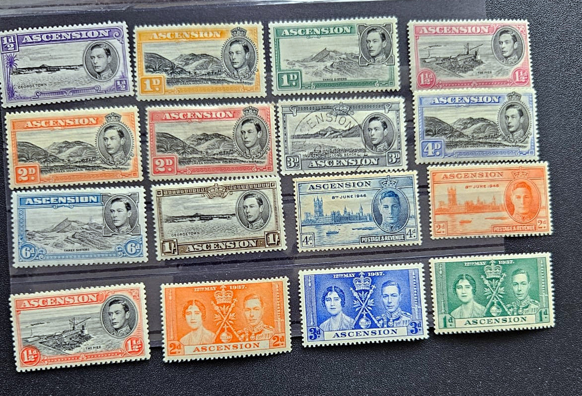 A set of 15 vintage Ascension Island postage stamps from the British Empire, arranged in a 3x5 grid on a black background. The stamps feature various colors including purple, orange, green, blue, and gray, with portraits of King George V and Queen Elizabeth and depictions of ships and landscapes.