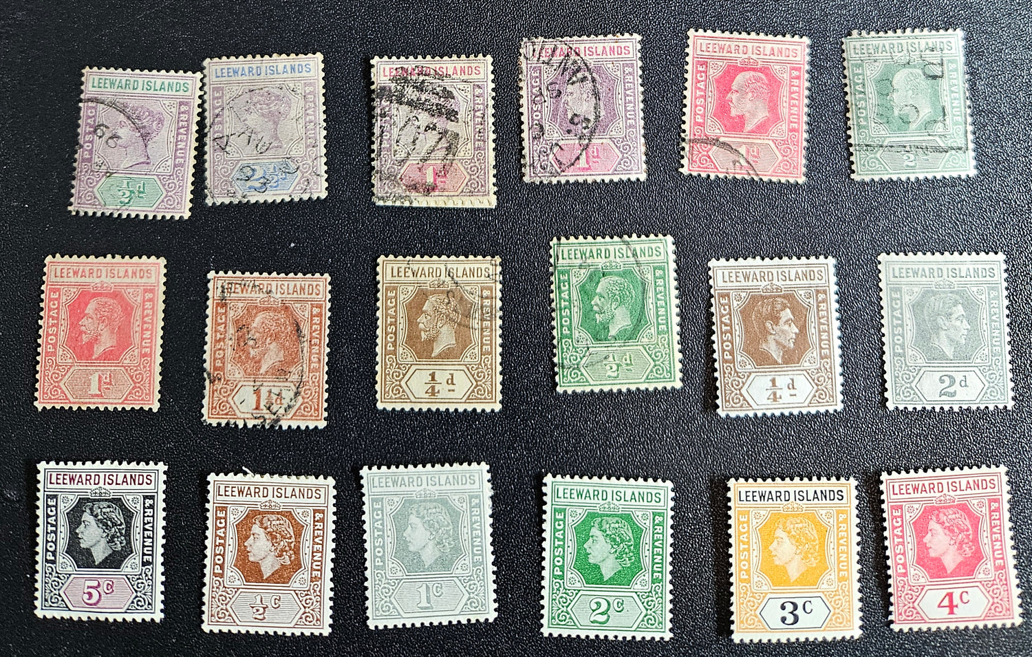 A collection of vintage Leeward Islands stamps featuring various monarchs from Queen Victoria to Queen Elizabeth II, displayed on a flat surface.