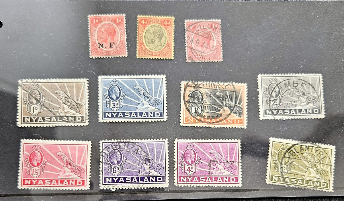 A set of vintage Nyasaland postage stamps arranged on a dark surface. The collection includes stamps in red, orange, pink, blue, gray, brown, purple, and olive-green colors, with some showing postmark cancellations and others featuring the word &