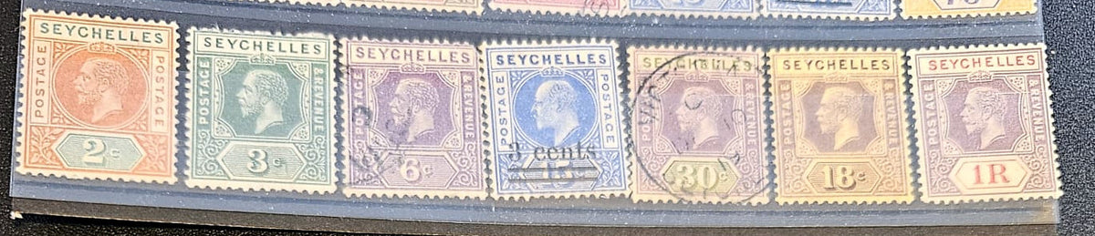 A close-up of a row of vintage Seychelles postage stamps in a clear plastic holder, showing multiple colors including orange, green, blue, purple, and yellow with profile portraits and denominations from 2 to 1R.