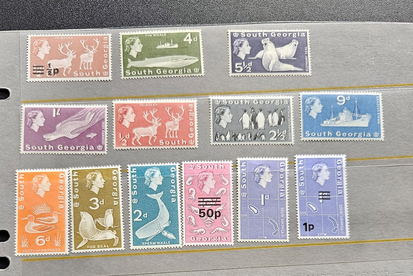 A set of vintage South Georgia postage stamps arranged on a gray binder page. The collection includes stamps in various colors such as green, purple, blue, pink, orange, and brown, featuring wildlife and landscape illustrations.